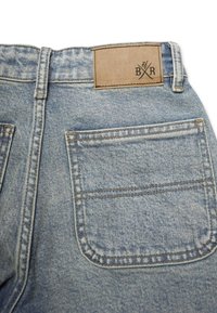 Vaaleansiniset farkut, joissa on nahkalaatta vyötäröllä ja yksi takatasku, jossa on vahvistettu ommel ja kulunut pinta.