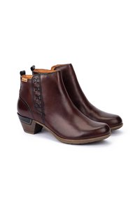 Par de botas de tobillo de cuero marrón con tacones bajos apilados, costuras decorativas y lengüetas en un fondo blanco.