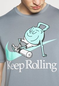 Camiseta gris con un gráfico divertido de un personaje relajándose sobre un rodillo de espuma, con acentos turquesa y un texto blanco en negrita que dice "Sigue Rodando".