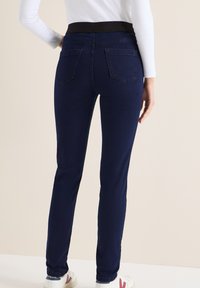 Donkerblauwe jeans met een taps toelopende pasvorm en elastische tailleband, voorzien van twee achterzakken en een gladde denimtextuur. Gestyled met witte schoenen.