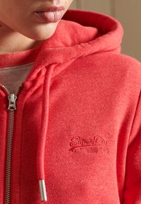 Primer plano de una persona que lleva una sudadera roja de SuperDry con cordones y cremallera de metal, mostrando parcialmente los labios y la barbilla.