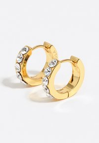 Pendientes de aro dorados que presentan una fila de rhinestones claros a lo largo de un lado, creando un diseño brillante y moderno. Superficie suave con un acabado pulido.