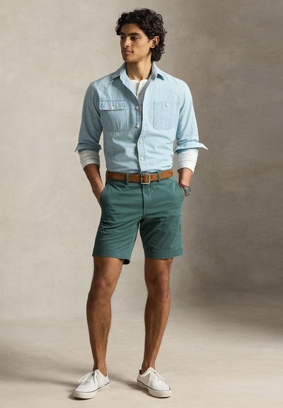 Tânăr stând cu mâinile în buzunare, purtând o cămașă din denim albastră peste un tricou alb cu mâneci lungi, pantaloni scurți verzi, curea maro și adidași albi.