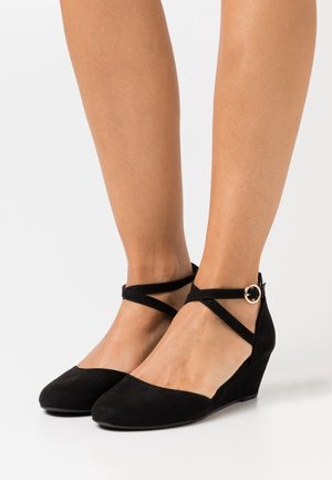 Jambes portant des talons compensés en daim noir à bouts pointus avec des brides croisées autour de la cheville, maintenues par une petite boucle dorée ronde.