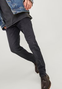Tmavě šedé slim-fit džíny s hladkou texturou, kombinované s hnědými šněrovacími botami a denimovou bundou nošenou přes vrstvený tmavý top.
