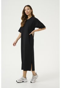 Black, knee-length dress made of soft fabric, featuring short sleeves and side slits. Paired with gray and white athletic shoes.