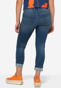 Sininen denim-materiaaliin perustuvat slim fit -farkut, joissa on käännetyt lahkeet, takataskut ja kontrastivärinen ompelu, yhdistettynä oransseihin tennareihin neutraalilla taustalla.