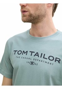 Hellgrünes Baumwoll-T-Shirt mit Rundhalsausschnitt. Große schwarze Schriftzüge "TOM TAILOR" und "THE CASUAL DEPARTMENT" auf der Vorderseite.