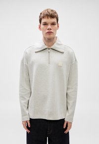 Ανοι χτό γκρι πουλόβερ με ψηλό γιακά, μερικό φερμουάρ, υφασμάτινη υφή και τετράγωνο λογότυπο στο στήθος. Μακριά μανίκια, άνετη εφαρμογή.