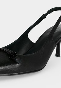 Chaussure à talon haut en cuir noir avec bride arrière, bout pointu et petite boucle décorative à l'avant.