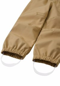 Tannfarbene, wasserfeste Hose mit elastischen Bündchen, ausgestattet mit weißen Schlaufen an den Knöcheln, zwei Taschen an den Seiten und einer glatten, glänzenden Textur.