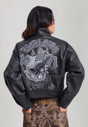 Femme aux cheveux foncés porte une veste en cuir noire avec un dessin d’aigle et du drapeau américain et le texte "Ed Hardy 1971 Born Free New York" dans le dos.