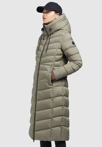 Long olive green puffer coat with a high collar, horizontal stitching, side pockets, and a zipper closure. Paired with a black cap.