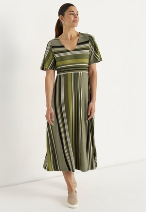 Frau mit dunklem Haar im Pferdeschwanz trägt ein grün gestreiftes Midikleid und beige Slip-On Schuhe und geht vor einem schlichten weißen Hintergrund.