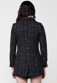 Blazer texturat negru cu model în carouri albe, având un guler în formă de V, mâneci lungi și tiv franjurat. Se potrivește deasupra coapselor.