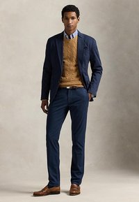 Jonge man staat met hand in zak, draagt een marineblauw colbert, een beige kabelgebreide trui, blauwe broek en bruine loafers.