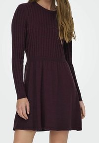 Femme portant une robe à manches longues en maille côtelée de couleur violet foncé, avec une taille cintrée et une jupe évasée, sur un fond uni.