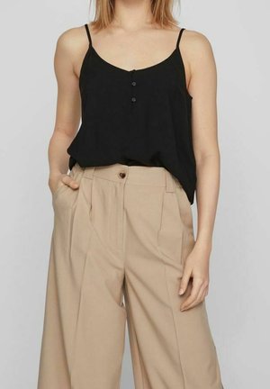 Woman wearing a black sleeveless buttoned top tucked into high-waisted wide-leg beige trousers, one hand in pocket.