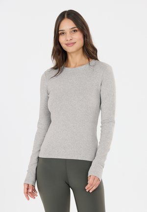 Kvinde med langt brunt hår iført en tætsiddende lysegrå langærmet trøje og mørkegrønne leggings, smilende med en ensfarvet hvid baggrund.