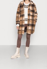 Personne portant un manteau épais à carreaux beige et marron, un pull blanc à motifs, un pantalon marron et des bottes crème épaisses, debout devant un fond blanc.