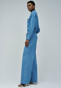 Salopette en denim bleu à coupe décontractée, dotée de manches longues, d'une poche poitrine et de jambes larges. Texture lisse avec un design classique.