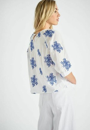 Witte blouse met blauw bloemmotief, korte mouwen, ronde halslijn met sleutelgatdetail aan de achterkant, gecombineerd met lichtgekleurde broek.