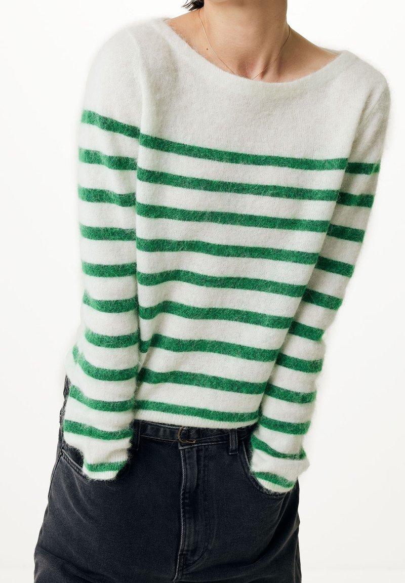 Pull blanc duveteux avec des rayures horizontales vertes uniformément espacées, porté avec un pantalon sombre taille haute, mains dans les poches, sur fond uni.