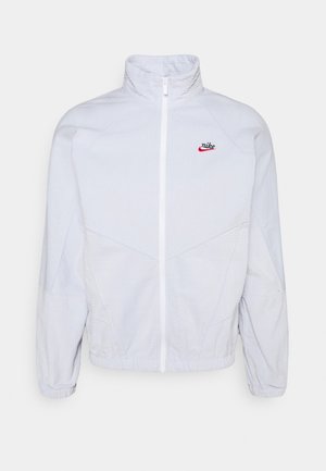 Witte Nike-jas met volledige ritssluiting, hoge kraag, lange mouwen, elastische manchetten en een klein rood-zwart Nike-logo op de borst.