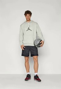Grå hoodie med en centrerad svart logotyp, i kombination med svarta mönstrade shorts. Modellen håller en basketboll och har på sig sneakers med röda accenter.