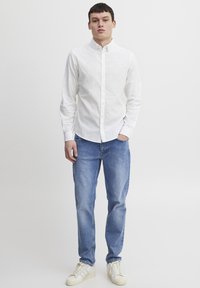 Camisa blanca de manga larga abotonada hecha de lino, combinada con vaqueros de denim azul claros y zapatillas blancas con detalles grises.