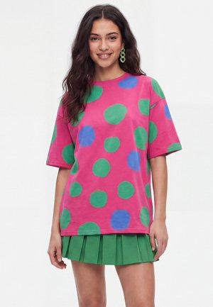 Ung kvinde smiler iført en pink oversized skjorte med grønne og blå polkaprikker, et grønt plisseret miniskørt og øreringe.