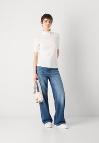 Hvit kortermet topp med mock-neck, parret med blå vide jeans. Holder en liten beige veske, komplementert av hvite sneakers.