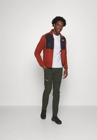 Röd och svart zip-up jacka över en vit t-shirt; gröna cargobyxor med sidofickor; svarta sneakers. Neutralt bakgrund.