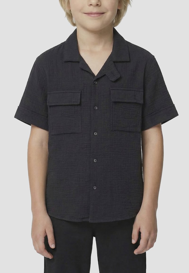 Enfant aux cheveux clairs porte une chemise noire à manches courtes boutonnée, avec deux poches poitrine et un col décontracté.