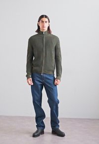 Olīvu zaļš ribotais džemperis ar rāvējslēdzēju, saskaņots ar ziliem džinsu biksēm un melniem potīšu zābakiem; modele stāv pret neitrālu fonu.