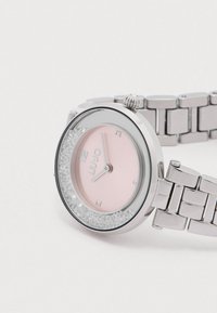 Ceas din metal argintiu cu o carcasă rotundă, față roz și accente cu cristale. Dispune de marcaje orare argintii și o curea de oțel inoxidabil.