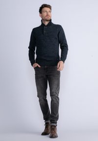 Maglione di cotone blu scuro con colletto alto e due bottoni. Indossato con jeans grigi slim-fit e stivali stringati marroni. Design semplice e con texture.