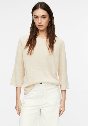 Młoda kobieta z długimi brązowymi włosami, ubrana w luźny beżowy sweter z rękawami do łokci i białe spodnie z wysokim stanem, stojąca na białym tle.