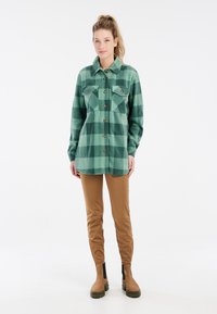 Groene geruite flanellen overhemd met metalen knopen, kraag en twee voorzakken. Gecombineerd met bruine fitted broeken en bruine enkelboots.
