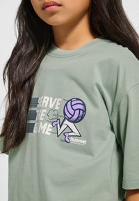 Grön bomull t-shirt med kort ärm som har en grafisk bild av en löpande figur och volleyboll, texten "Serve the Game" och varumärkeslogotyp.