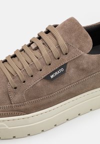 Brązowe zamszowe sneakersy z beżowymi sznurówkami, z gumową podeszwą i przeszytymi akcentami. Z boku widoczna jest czarna metka z napisem "MORATO".