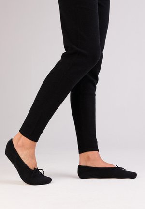 Ballerines noires en tissu doux, avec un bout rond et un petit nœud sur le devant, portées avec un pantalon noir ajusté.