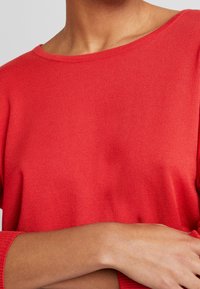 Roter gerippter Pullover mit weitem Ausschnitt und langen Ärmeln, der eine glatte Textur und eine taillierte Silhouette präsentiert.
