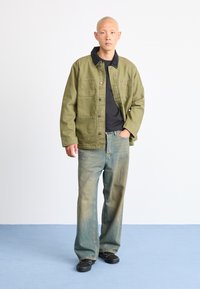 Olivengrøn jakke med patchlommer, sort krave, parret med falmede blå vidde jeans og sorte sko på et blåt gulv.