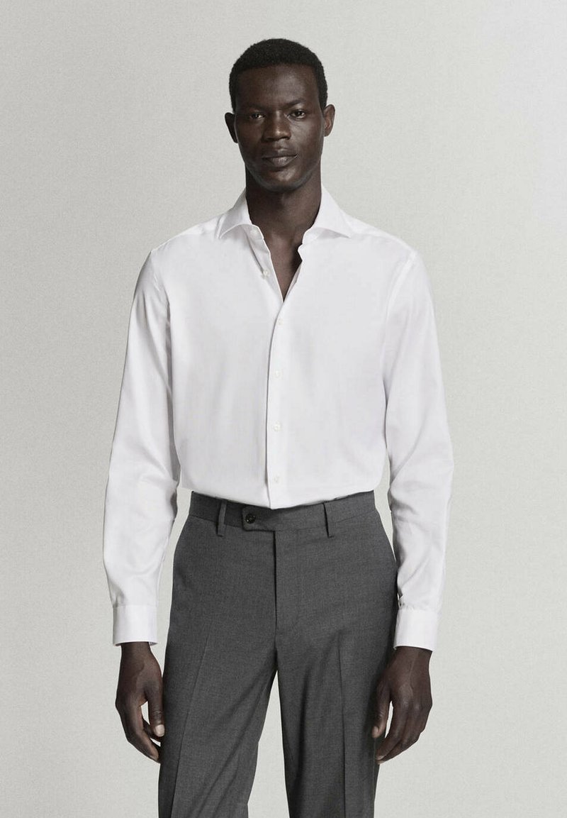 Homme debout face à l’objectif, portant une chemise blanche à manches longues boutonnée et un pantalon gris sur un fond uni.