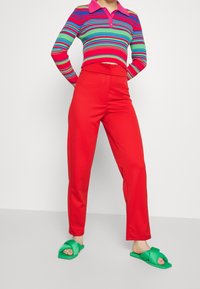 Personne portant un pantalon taille haute rouge, une chemise à rayures colorées à manches longues, et des sandales ouvertes vertes se tenant devant un mur blanc.