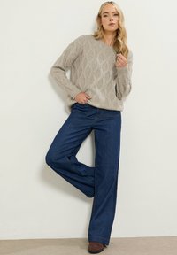 Beige kabelstrikket sweater med lange ærmer, parret med højtaljede, brede mørkeblå denim jeans. Brune sko synlige. Neutral baggrund.