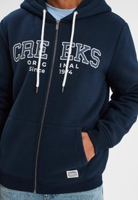 Sudadera con cremallera en color marino, hecha de algodón, con letras blancas, cordones y un bolsillo canguro. Incluye una etiqueta de la marca en el dobladillo.