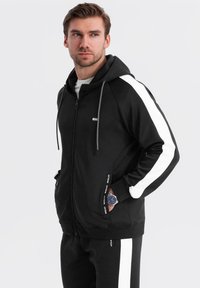 Uomo che indossa una felpa nera con zip e strisce bianche sulle maniche e pantaloni abbinati con le mani nelle tasche, su sfondo bianco.