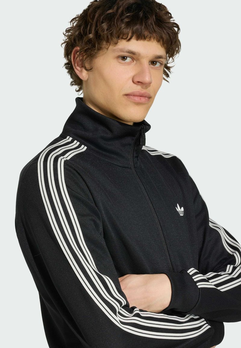Junger Mann mit kurzen lockigen Haaren, der eine schwarze Adidas Trainingsjacke mit weißen drei Streifen trägt und die Arme verschränkt hat.